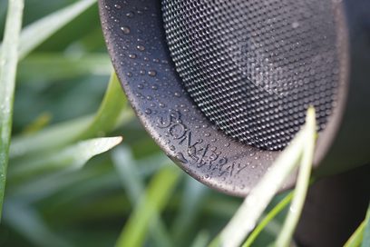 Sort perforeret metalrist til højttaler med indskriften "Sonarray" og vanddråber på uskarp grøn bladbaggrund