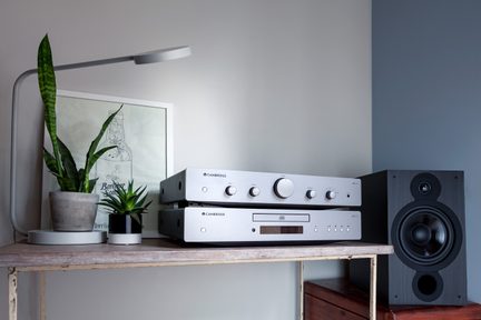 Silver metal hi-fi system viewed from the front, consisting of a stacked amplifier and CD player with buttons and a large volume knob, placed on a wooden shelf next to a black wooden speaker, under a gray metal lamp and between two potted plants.