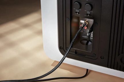 Close-up rear view of a subwoofer, glossy white rectangular enclosure, black plastic connection panel with two notched knobs, audio jacks, and a connected black cable