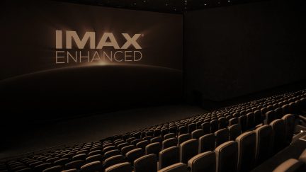 Sala cinematografica vuota, sedili allineati, schermo gigante con testo "IMAX ENHANCED", vista angolata, illuminazione soffusa