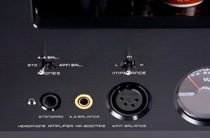 Vista ravvicinata frontale di un pannello anteriore in alluminio spazzolato nero di un amplificatore per cuffie, con due interruttori a levetta argentati, un jack da 6,35 mm nero, un jack bilanciato da 4,4 mm con bordo dorato, una presa XLR a 4 poli nera e un VU-meter parzialmente visibile