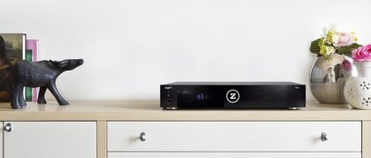 Zappiti Neo multimedie-afspiller, set forfra, blå LED-display "155:1", rektangulært sort kabinet, på lyst trækabinet