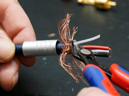Primo piano obliquo su un cavo audio con guaina in plastica blu dotato di un manicotto cilindrico metallico argentato, il cui schermo intrecciato in rame è scostato per mostrare i conduttori rosso, nero e bianco, tenuto tra due dita e un tronchese nero con impugnature rosse e blu.