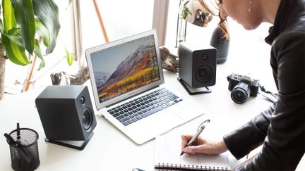 Mustat työpöytäkaiuttimet avoimen MacBook Airin molemmin puolin valkoisella pöydällä kameran kanssa