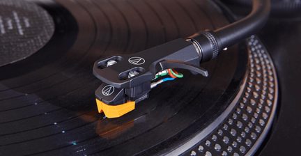 Nahaufnahme der schwarzen Phonozelle aus Kunststoff und Metall mit orangener Spitze und sichtbaren farbigen Kabeln, schräge Ansicht auf eine schwarze Schallplatte, die auf einem metallisierten Plattenteller liegt