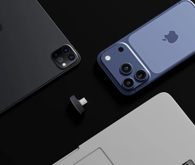 Adaptor USB-C negru înconjurat de un iPhone albastru, un iPad gri și un laptop argintiu pe fundal negru