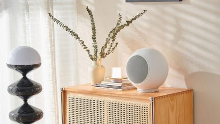Composizione decorativa d’interni, vista a tre quarti all’altezza degli occhi, che mostra su una credenza in legno chiaro traforato un diffusore sferico bianco in plastica opaca con griglia metallica frontale, affiancato da un vaso in ceramica beige contenente rami, una piccola candela e, a sinistra, una lampada scultorea nera laccata con cupola bianca