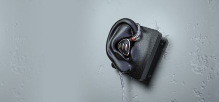 Schwarzer In-Ear-Kopfhörer mit dreieckigem Metallgehäuse und kupferfarbenem Rand, zwei dekorativen Schlitzen und durchsichtig geflochtenem Kabel, eingesetzt in ein schwarzes Silikon-Ohrmodell, das auf einer rechteckigen Halterung befestigt ist, leicht schräg von vorne gesehen