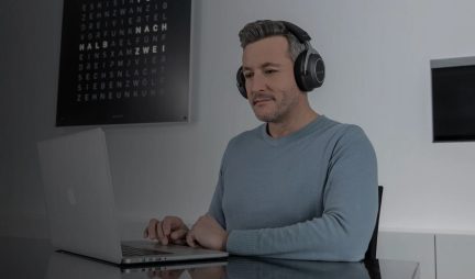 Άνδρας καθιστός, μαύρα ακουστικά, μπλε πουλόβερ, χρησιμοποιώντας ασημένιο φορητό υπολογιστή, μαυροπίνακας με κείμενο στο φόντο