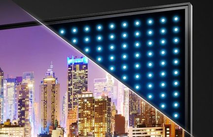 Esquina superior derecha de una pantalla plana, marco negro con acabado metálico visto en tres cuartos, corte diagonal que revela una retroiluminación interna en matriz regular de LEDs azules detrás de una imagen luminosa de rascacielos nocturnos.
