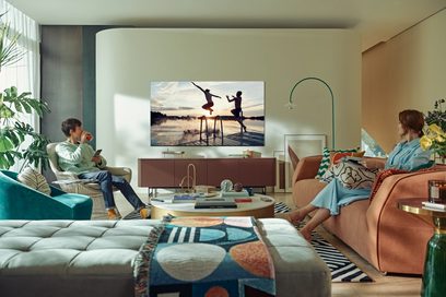 Moderne stue med to børn siddende på blå sofaer og ser fjernsyn, hvor to personer hopper på en bådebro ved solnedgang
