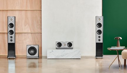 Frontvisning af et sæt hi-fi-højttalere bestående af to gulvhøjttalere, en subwoofer og en centerhøjttaler, rektangulære kabinetter i grå aluminium og sort højglans med synlige runde højttalerenheder, placeret i et moderne interiør med poleret gulv