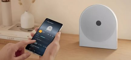 Altoparlante smart grigio dal design essenziale su un tavolo in legno, controllato da uno smartphone in primo piano