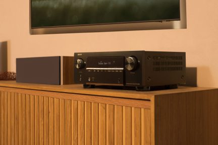Černý AV receiver Denon pro domácí kino s velkým středovým displejem a dvojitými otočnými ovladači, pohled ze tří čtvrtin na dřevěném nábytku