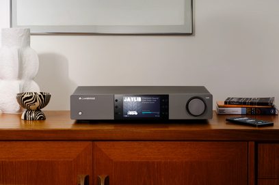 Síťový audio přehrávač EXN100 v černé barvě, pohled zepředu, centrální displej rozsvícený, kruhové tlačítko vpravo, umístěný na dřevěném nábytku