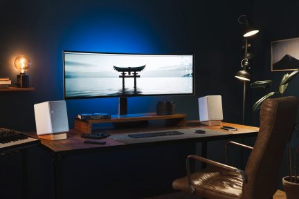 Houten bureau met gebogen scherm, toetsenbord, muis, witte luidsprekers, lamp, leren stoel, plant, plank en blauwe muur