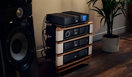 rack hi-fi cu patru niveluri, rafturi din lemn deschis la culoare, montanți din metal alb, module audio din metal negru, fațade netede, buton rotativ circular iluminat, ecran pătrat albastru, vedere din unghi de trei sferturi