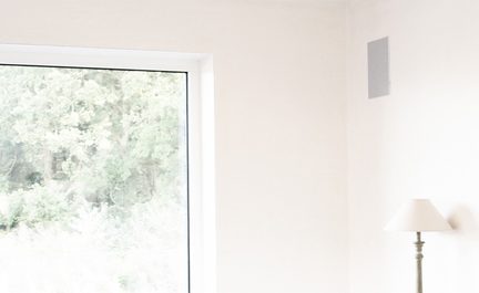 Pilkinė į sieną įmontuojama kolonėlė šviesiame svetainės kambaryje su dideliu langu ir balta toršere