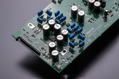 Zelený tištěný spoj s modrými a černými kondenzátory, rezistory a dalšími elektronickými součástkami, detailní pohled shora