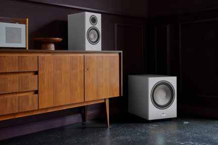 valkoinen kirjahyllykaiutinsarja ja vaaleanharmaa subwoofer matalan vintage-tyylisen puisen senkin vieressä olohuoneessa