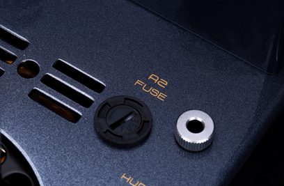 Primer plano oblicuo del panel superior metálico negro texturizado del amplificador a válvulas Cayin CS-805A, que muestra ranuras de ventilación, la inscripción naranja «A2 FUSE», un tapón de fusible circular negro y un inserto roscado plateado.