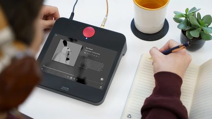 Pohled shora, černá obrazovka s červeným tlačítkem, ruka píšící do notesu, bílý hrnek, zelená rostlina v květináči