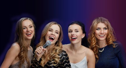 Gruppe af fire unge kvinder i aftenkjole, der synger karaoke med mikrofon på en lilla-blå tonet baggrund