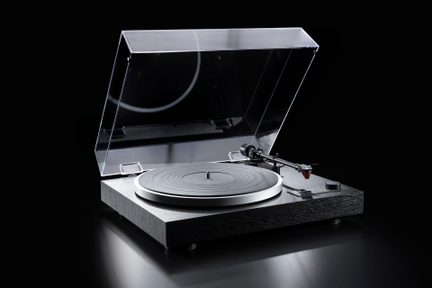 Svart vinylspelare med träbas i textur, matt grått räfflat cirkulärt tallrik, svart metallisk tonarm med röd pickup, uppfällt transparent dammskydd, vy snett framifrån