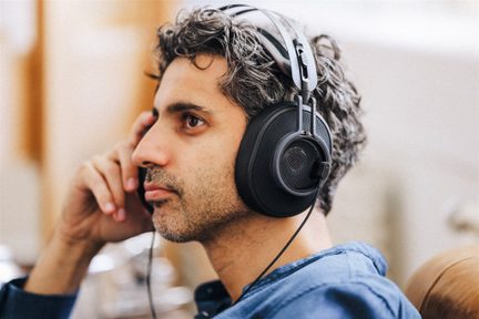 Prim-plan din profil al unei căști audio cu fir, de culoare neagră, din plastic și metal, cu cupe circumaurale cu grilaj din plasă și bandă dublă, purtată de o persoană