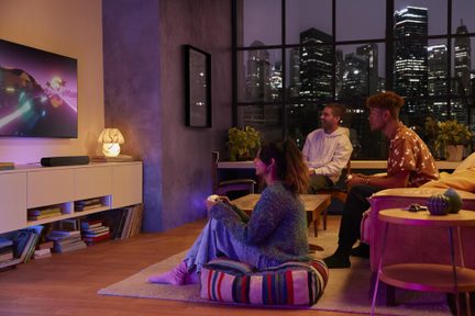 Soundbar nera sotto TV da gaming, soggiorno moderno con tre amici che giocano, illuminazione ambientale colorata e grande vetrata sulla città