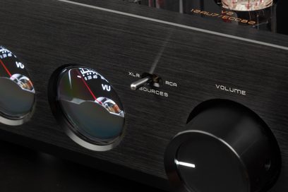 Close-up, slightly top-down front view of the Cayin HA-3A high-fidelity headphone preamplifier’s black brushed aluminum front panel, featuring two domed VU meters, a central XLR/RCA lever selector, and a large volume knob on the right