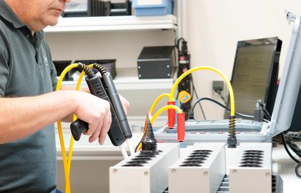Detailní záběr ruky technika držícího dvě černé plastové sondy-pistole spojené žlutými spirálovými kabely před modulárním kovovým testovacím stolem s válcovými černými výřezy, pohled ze tří čtvrtin