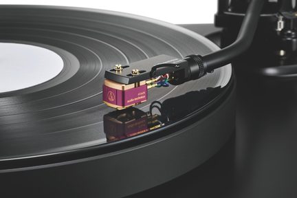 Skrå visning af en sort vinylplade på pladespiller, metal-tonearm, magenta og gylden pickup, flerfarvede kabler