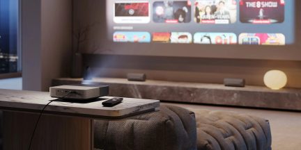 Videoproiettore compatto nero con telecomando su tavolino di fronte a uno schermo di streaming in un soggiorno moderno