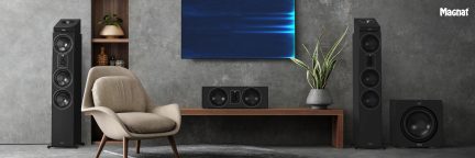 Woonkamer met grijze muur, beige fauteuil, houten tafel, 2 zwarte zuilluidsprekers, 1 centerspeaker, 1 subwoofer, groene plant, wandgemonteerd scherm