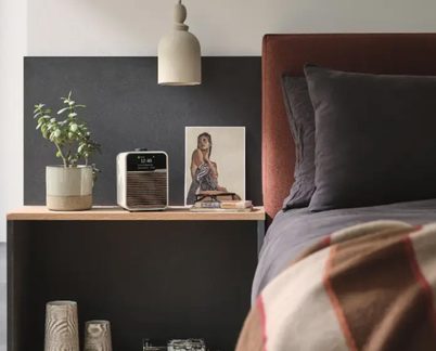 Puinen yöpöytä, beige keramiikkalamppu, kasviruukku, musta ja hopeanvärinen herätyskello, ruskea sängynpääty, harmaat lakanat