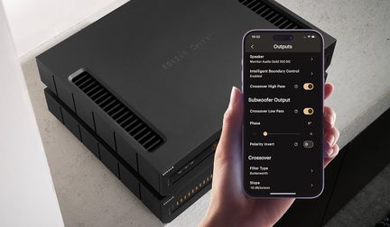 Svart forsterker stablet, sett ovenfra, hånd som holder smarttelefon som viser lydinnstillinger, grå overflate i bakgrunnen