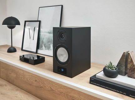 Černá hi-fi regálová reprosoustava s dvojicí kulatých reproduktorů, z nichž jeden má světlý kužel, umístěná na moderním dřevěném nábytku