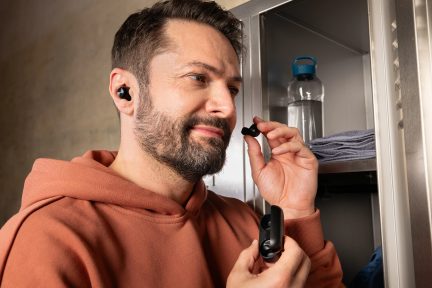 Brodaty mężczyzna w pomarańczowej bluzie z kapturem trzymający czarne bezprzewodowe słuchawki i etui przed otwartą metalową szafką