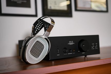 Cuffie argentate con archetto perforato su amplificatore nero, pulsante circolare, prese audio, superficie in legno, vista laterale