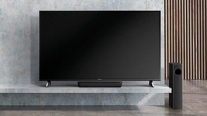 Sort Panasonic SC-HTB250EGK soundbar placeret på en lys grå møbel foran et stort fladskærms-TV med sort skærm, der er slukket