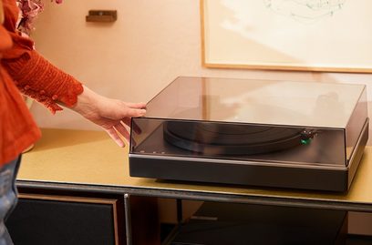 Cambridge Audio Alva ST hõbedane vinüülmängija, kaas suletud, toonari kaar paigas, naiskäsi puudutamas plaaditaldriku