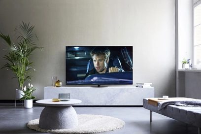 Téléviseur Panasonic TX-55HZ1000E allumé sur meuble blanc, affichant gros plan visage homme dans voiture, plante verte à côté