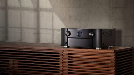 Amplificateur audio en métal brossé noir, deux gros boutons, écran rond central à halo bleu, vue 3/4 avant droit