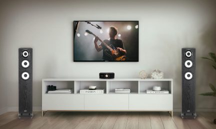 A wall-mounted TV displaying a musician, two black speakers, a white cabinet with decorative objects and books