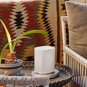 Trådløs, tilsluttet Sonos One (Gen 2) højttaler i mat hvid på et etnisk tæppe, med en grøn plante, puder og en fletstoflænestol