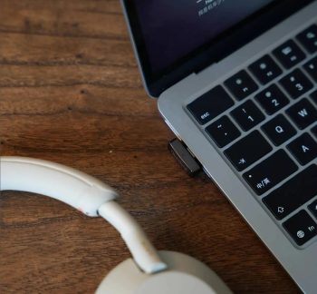 Adaptor USB negru conectat la un laptop gri, lângă o pereche de căști albe pe o masă maro