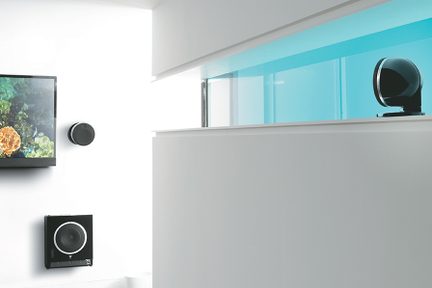 Perspektivisk udsigt over et hvidt, minimalistisk interiør med sort fladskærms-tv på væggen, ledsaget af en mat sort, cirkulær væghøjttaler, en blank sort, firkantet subwoofer og en sort, lakeret, sfærisk højttaler placeret bag en lyseblå glasvæg.