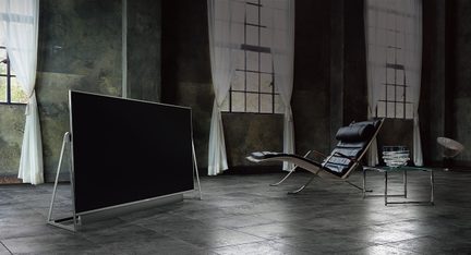 Fladskærms-tv på metalfod, lædersofa med sort læder der kan vippes og glasbordplade i et rum med nedbrudte vægge og hvide gardiner