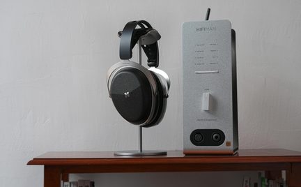 Sort over-ear hovedtelefon i sort og sølv på metalstativ, ved siden af en sølvfarvet hovedtelefonforstærker med drejeknap og stik, set forfra